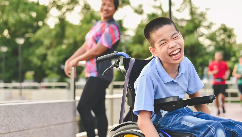 A boy in a wheelchair smiling with a smiling adult in the background